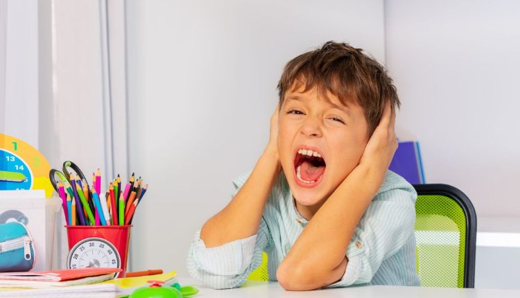 طفل يجلس على كرسي يغطي أذنيه ويصرخ من أحد مسببات الانهيار عند أطفال التوحد وأمامه طاولة وعلبة ألوان