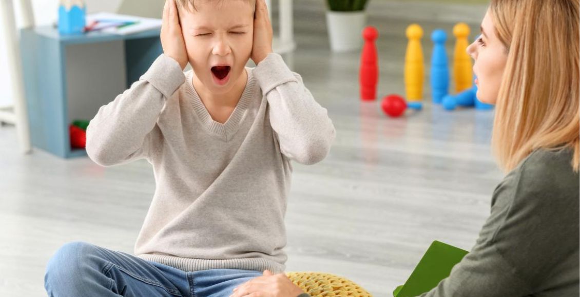 طفل يجلس في غرفة فيها ألعاب ويغطي أذنيه ويصرخ وبجانبه والدته تحاول معرفة كيفية تهدئة طفل التوحد