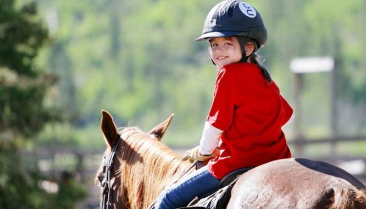 طفلة ترتدي قبعة وبلوزة حمراء وبنطال جينز وتمتطي خيل لتوضيح أهمية ركوب الخيل في علاج التوحد