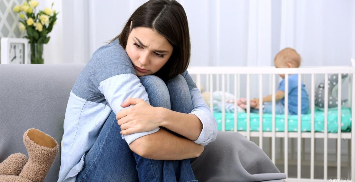 أم تجلس على الأرض حزينة تعاني من قلق ما بعد الولادة Postpartum Anxiety وبجانبها طفلها على السرير