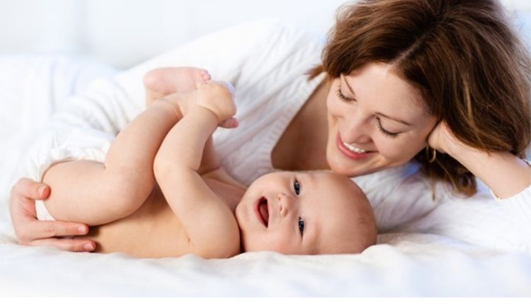 أم مبتسمة تداعب طفلها الرضيع على السرير لتوضيح أهمية معالجة قلق ما بعد الولادة