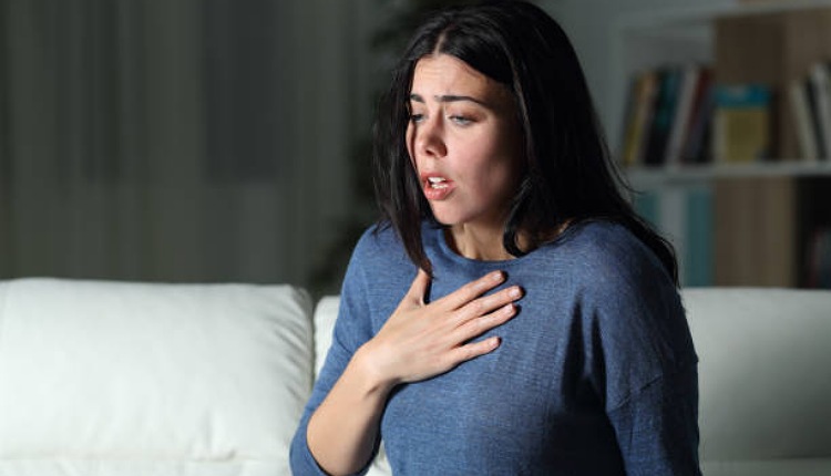 امرأة تجلس على أريكة بيضاء تضع يدها على قلبها تعاني من نوبة الهلع الخلفية رمادية